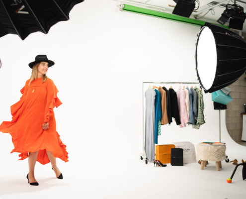 Woman on set in orange flowy dress with a black hat in front of white cyc wall next to clothing rack